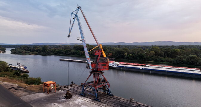 Aerial view of the Danube Port featuring a large industrial crane on the riverbank and cargo barges moored on the water with lush green trees in Pancevo, Serbia.