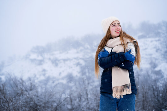 Cold young woman wearing warm casual winter clothing and trying to stay warm during snowfall outdoors. Winter weather and freezing temperature concept.