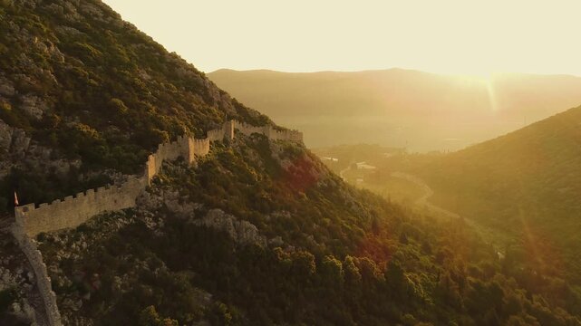 Aerial view of the historic stone fortifications of the Walls of Ston winding along the mountainous landscape during golden hour in Croatia.