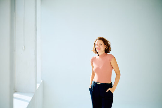 Professional woman smiling, standing with hands in pockets against a bright studio wall, projecting confidence and success