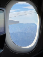 Fototapeta premium Airplane wing flying above rugged mountains with blue sky seen from passenger window
