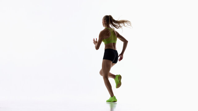 Rear view of runner gaining speed across clean white studio. Concept of forward momentum, endurance training and determined pursuit of personal goals.