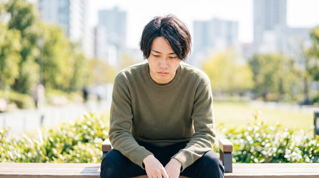 公園のベンチで落ち込んでうつむく悩みを抱えた若い男性