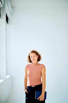 Young professional woman standing confidently, holding a blue notebook, in a bright modern office building interior