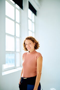 Mid aged professional woman with short curly hair smiling confidently, holding a folder in a bright modern office by large window