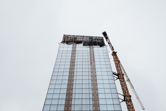A crane is positioned next to a tall building under construction with glass panels. The structure displays vertical lines and is surrounded by a gray sky