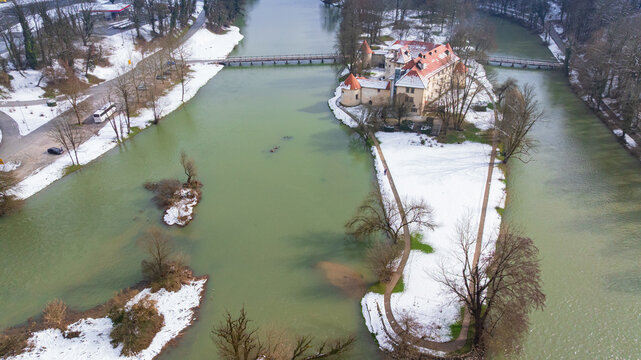 Picturesque drone panorama shows historic Otocec Castle covered in light snow. Frozen park trees, wooden bridges, and cold green Krka river frame medieval island palace in winter time.