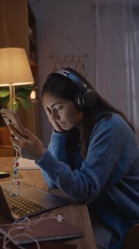 Vertical video of young adult Caucasian woman wearing headphones, rubbing her eyes in exhaustion while holding smartphone at desk. Ideal for mental health and digital wellness campaigns
