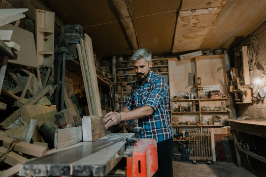 Carpenter working at a table saw in a cluttered woodworking workshop concentrating on cutting wood