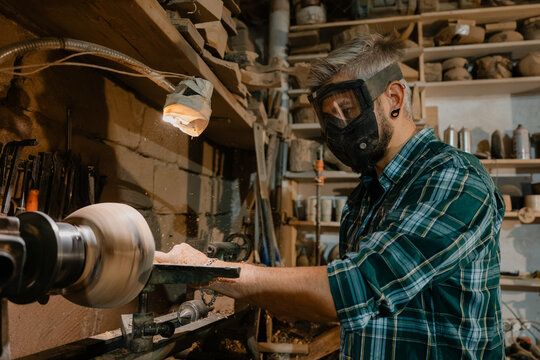 Woodworker shaping a spinning wooden bowl on a lathe in a dusty workshop wearing safety gear