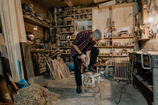 Experienced woodworker drilling a wooden stool using an electric drill in a cluttered rustic carpentry workshop