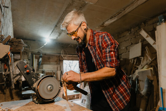 Man sharpening a blade on a bench grinder in a rustic home workshop wearing safety glasses