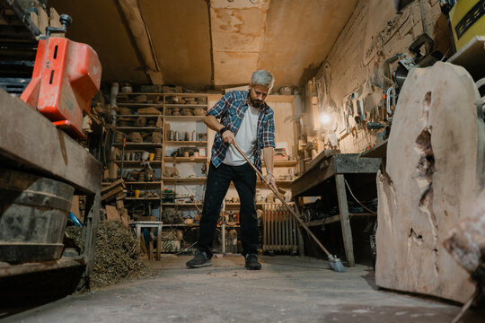 Middle-aged woodworker cleaning rustic workshop floor after a day of woodworking projects