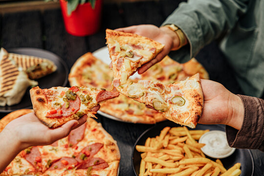 Close-up of several diners raising assorted pizza slices over plates of pizza, French fries, creamy dip and a grilled wrap at an outdoor restaurant table