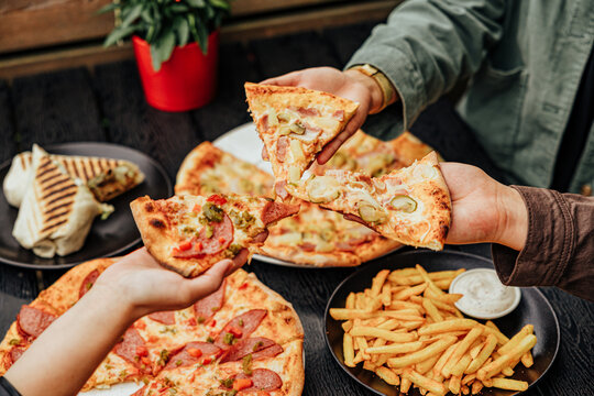 Close-up of several diners raising assorted pizza slices over plates of pizza, French fries, creamy dip and a grilled wrap at an outdoor restaurant table