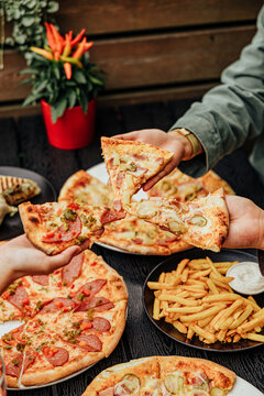 Close-up of several diners raising assorted pizza slices over plates of pizza, French fries, creamy dip and a grilled wrap at an outdoor restaurant table