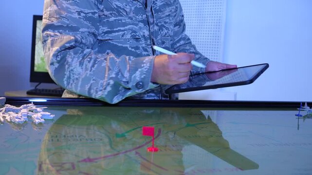 Military officer uses a tablet and stylus above an interactive digital map with a red location marker in a command center.