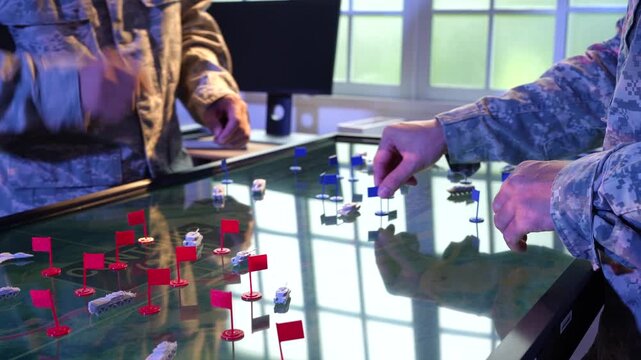 Military personnel in camouflage uniforms move pieces and red flags across a digital tabletop map during a tactical planning exercise.