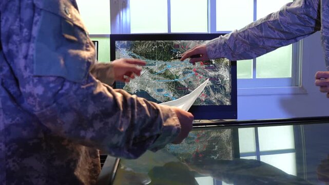 Military personnel in camouflage uniforms review a digital map on a monitor and touch table during a tactical planning meeting.
