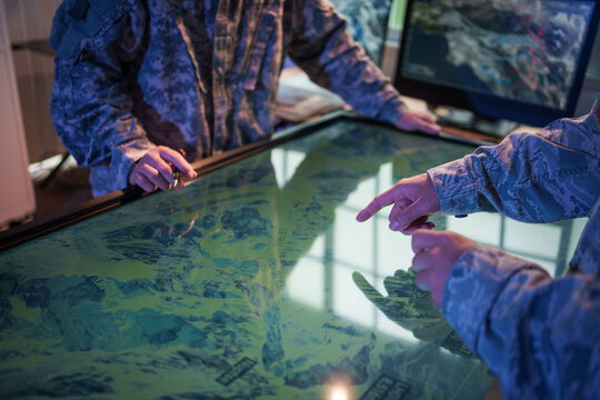 Military personnel in uniform use a large interactive map table for mission planning and tactical coordination in a command center.