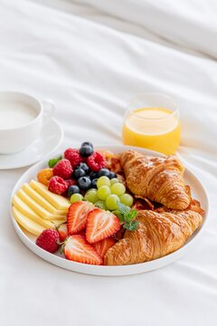 Cozy breakfast in bed with croissants, fresh berries, grapes, cheese, coffee and orange juice on white bedding