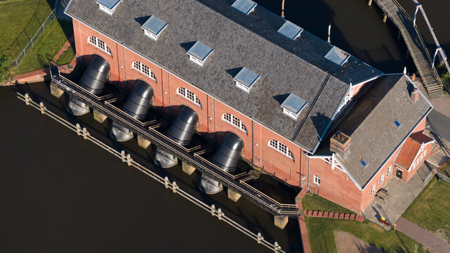 Aerial view of Gemaal De Waterwolf, a red-brick pumping station with large black discharge pipes extending into the water, in Lauwerzijl, Groningen, Netherlands.