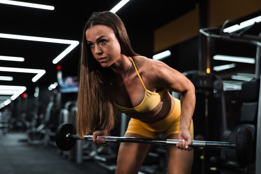 Yellow sportswear-clad woman focused on bent-over barbell row exercise in modern gym