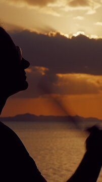 Vertical video. Silhouette of Woman Cooling Herself With Hand Fan During Golden Sunset by the Sea, Warm Evening Coastal Atmosphere, Relaxation, Summer Heat