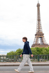 A young man walks in profile along a bridge near the Eiffel Tower in Paris, France. The lattice tower stands prominent in the background against a bright sky. © dsbedarieva