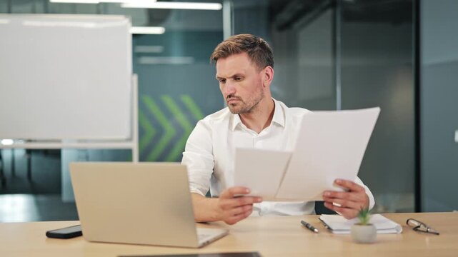 Professional reviews important business documents, cross-referencing information with laptop. Concentrated work in modern office setting. Focus on data analysis, problem-solving