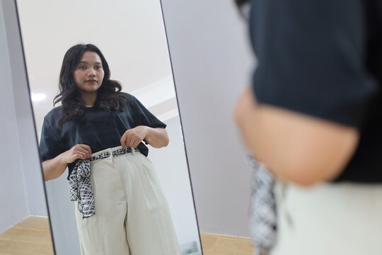A young woman stands in front of the mirror at home, adjusting her clothes and trousers, getting ready for work.