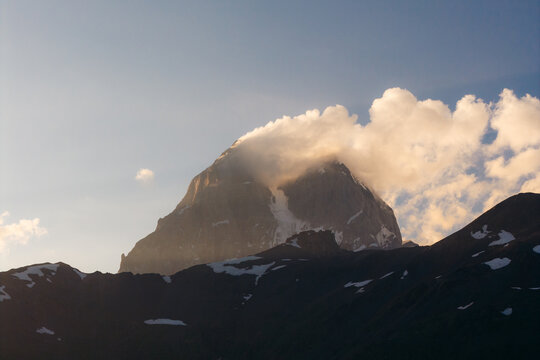 Aerial view of the snow-capped peak of Mount Ushba partially covered by clouds during golden hour in the Caucasus Mountains near Mestia, Samegrelo-Zemo Svanet, Georgia.