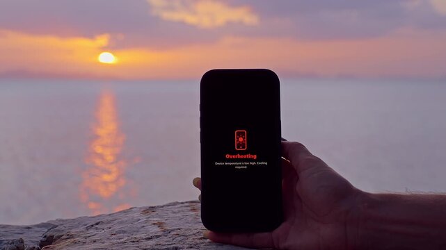 Hand Holding Smartphone Showing Overheating Warning Against a Sunset Sea Background, Mobile Device Safety, Heat Protection, Summer Technology Challenge