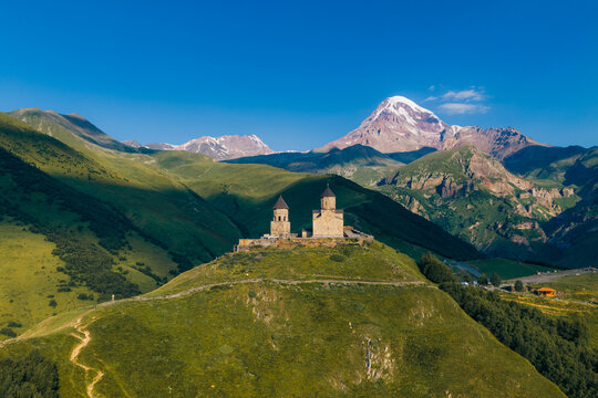 Aerial view of Gergeti Trinity Church perched on a grassy hill with the snow-capped Mount Kazbek in the background under a clear blue sky in Oni, Mtskheta-Mtianeti, Georgia.