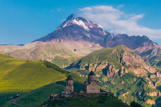 Aerial view of Gergeti Trinity Church perched on a hill against the backdrop of the snow-capped Mount Kazbek under a clear blue sky in Stepantsminda, Mtskheta-Mtianeti, Georgia.