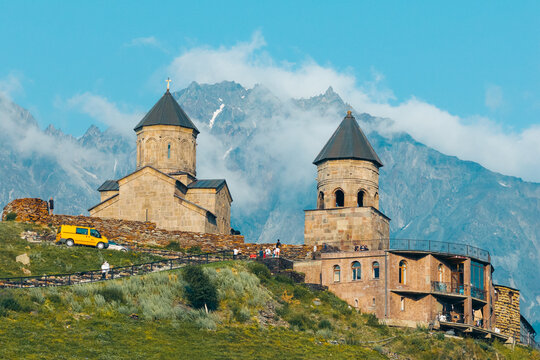 Aerial view of Gergeti Trinity Church set against the Caucasus Mountains with a yellow van and visitors on the historic grounds in Oni, Mtskheta-Mtianeti, Georgia.