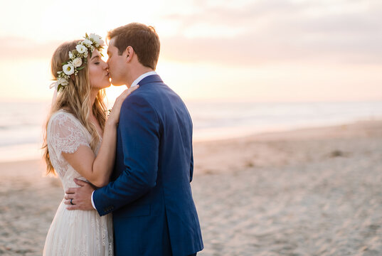 Young bride kissing groom on beach at sunset wearing white lace dress
