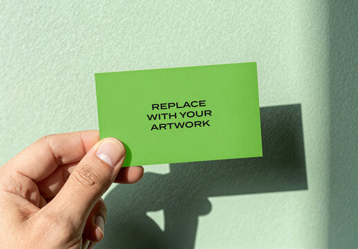 Business card mockup held in hand against a textured wall with natural light casting soft shadow, realistic stationery branding presentation, design