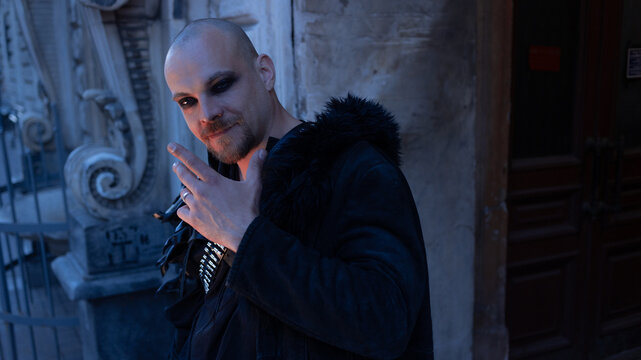 Man in gothic coat with raised hand and calm smile, welcome and invitation gesture.