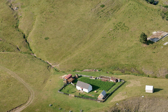 Aerial view of a small rural homestead with a white house, a garden, and an outbuilding situated in the green rolling hills of Montenegro.