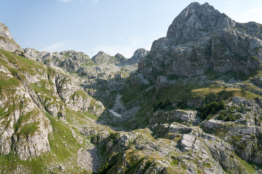 Aerial view of a rugged mountain range with rocky peaks, green slopes, and patches of snow in the valley under a clear sky in Montenegro.