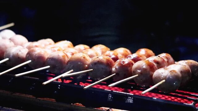 Thai street food vendor checks Isan sour sausage skewers over glowing charcoal at a night market, showing authentic local cuisine, hands on food service, and small business activity.