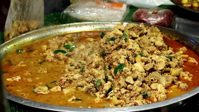 A street food vendor uses a large metal ladle to scoop steaming Thai red curry chicken with kaffir lime leaf from a steel tray into a plastic bag at a busy night market stall.