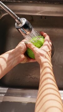Vertical video. Overhead View of Person Washing Green Avocado in Kitchen Sink in Slow Motion, Fresh Produce Hygiene and Healthy Nutrition