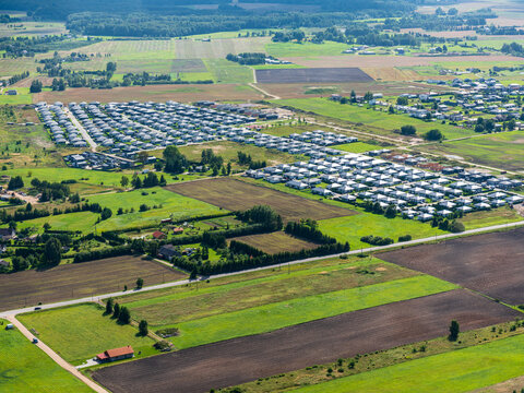 Aerial view of a modern residential neighborhood with rows of houses surrounded by vast agricultural fields, green meadows, and rural roads in Kaunas, Kaunas County, Lithuania.