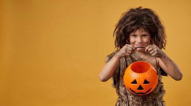 Smiling Boy Wearing a Prehistoric Caveman Halloween Costume with Brown Fur Outfit Holding an Empty Jack-o-Lantern Candy Bucket on an Orange Background