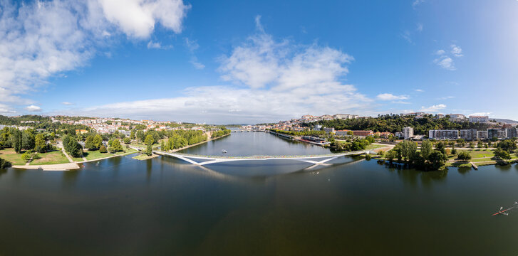 Aerial view of the modern Pedro e Ines pedestrian bridge spanning the Mondego River, surrounded by green riverbanks and the city skyline in Coimbra, Coimbra, Portugal.