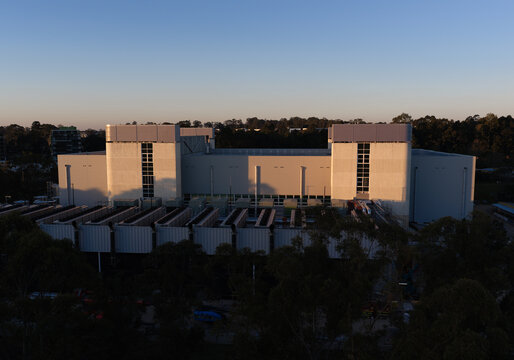 Aerial view of a modern data center facility with industrial cooling units on the roof, surrounded by lush trees under the soft light of the golden hour in Sydney, New South Wales, Australia.