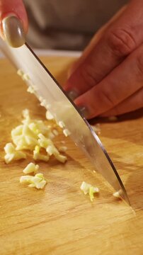 Vertical video. Close View of Female Hands Finely Chopping Fresh Garlic on Wooden Kitchen Board, Aromatic Seasoning and Homemade Food Preparation