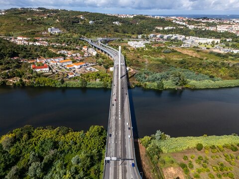 Aerial view of Ponte Rainha Santa Isabel spanning the Mondego River, surrounded by lush green hills, suburban buildings, and a clear blue sky in Coimbra, Coimbra, Portugal.
